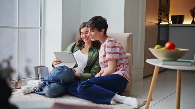 Happy sisters chatting online meeting talking to tablet web camera sitting floor