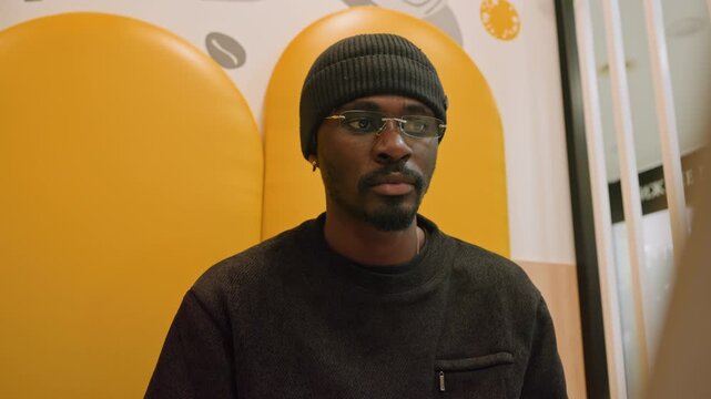 Man working laptop yellow booth focused on notes, pen in hand coffee cup nearby wearing beanie and glasses, warm yellow background urban cafe interior productive digital nomad aesthetic planning