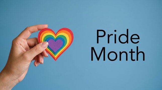Human hand holding a vibrant layered paper rainbow heart shape against a solid soft blue background representing equality love and inclusivity during the colorful global celebration of annual pride mo