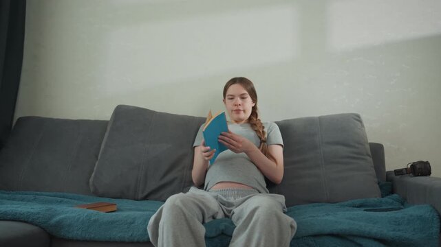 Pregnant caucasian woman on sofa reading guide, flipping teal manual pages, attentive expression, snack and phone nearby, casual grey tee and sweatpants, daylight fills quiet living room, researching