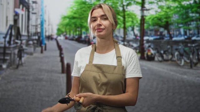 Woman gardener holding pruners with khaki apron and bare forearms visible on street;confidence readiness.