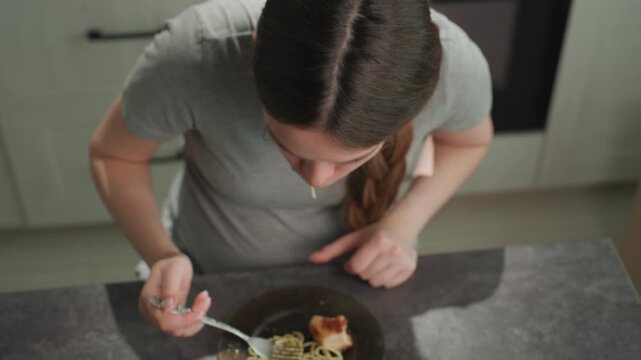 Young white woman eating spaghetti, twirling fork on kitchen counter, home cook takes bite then coughs and holds stomach indicating discomfort and nausea, braided hair and gray shirt, modern oven