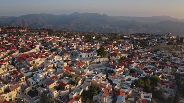 Pano Lefkaras traditional red tile roofs and narrow streets cascade down a hillside village in Cyprus, bathed in warm golden sunset against rolling mountains. Rural cityscape aerial view drone flight
