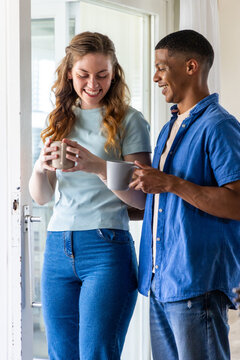 Diverse couple standing in bright home doorway holding ceramic mugs near glass door smiling