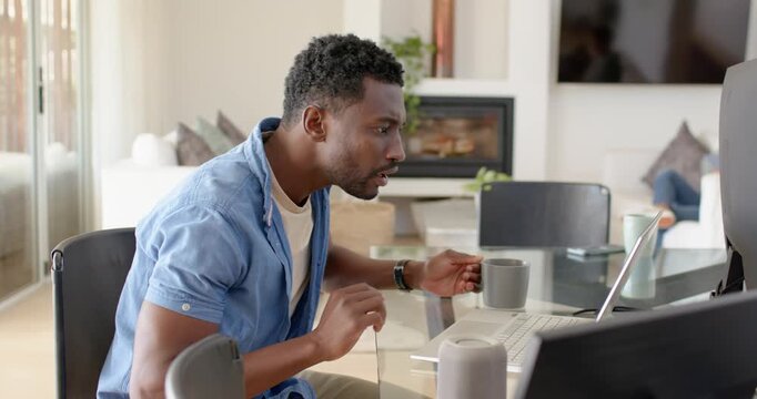 African American man in blue tee at home working, typing, clicking laptop, holding mug, celebrating
