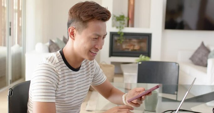 Asian man sitting at glass table, tapping pink phone, glancing at laptop, celebrating win