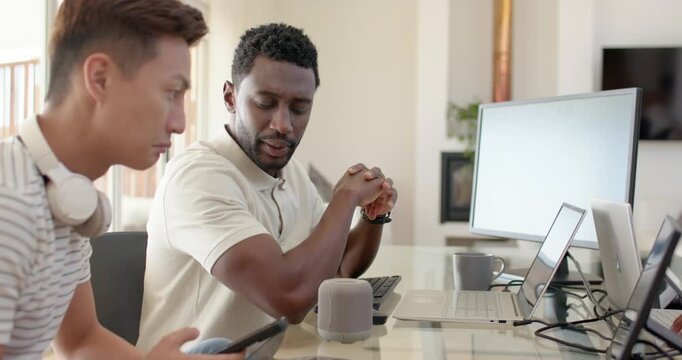Diverse men in home office, left demonstrating tablet, right testing speaker and typing on laptop