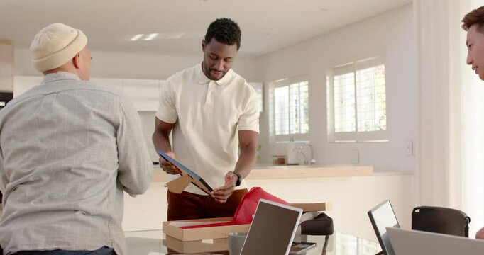 Diverse male friends in kitchen opening red bag, lifting framed plaque on glass table, celebrating