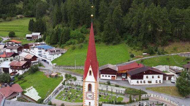 Aerial 4K drone shot of Nauders alpine village in Austria, featuring the iconic church steeple, traditional roofs, and scenic green mountain valley in the Tyrolean Alps on a cloudy day.