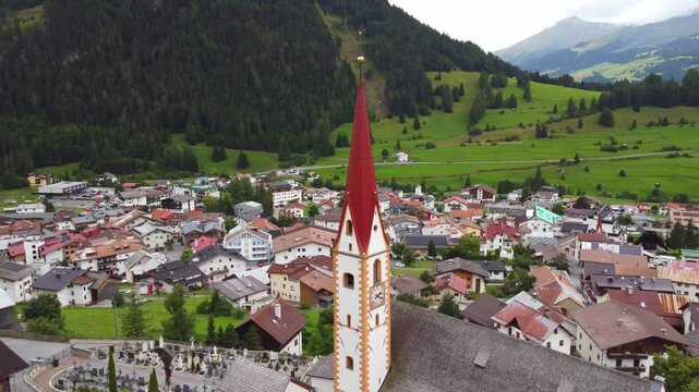 Aerial 4K drone shot of Nauders alpine village in Austria, featuring the iconic church steeple, traditional roofs, and scenic green mountain valley in the Tyrolean Alps on a cloudy day.
