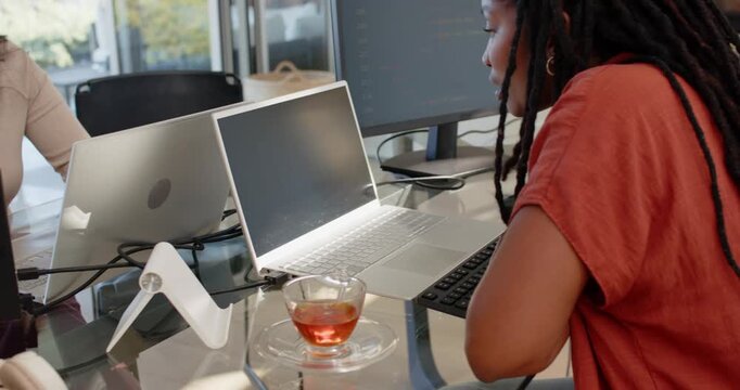 Diverse female coworkers at glass table one in beige top responding to monitor alert typing laptops