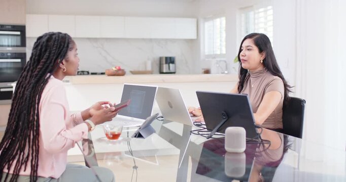 Diverse women working at glass table left showing phone right typing laptop third entering with box