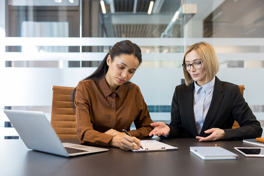 Two businesswomen collaborating on a project, reviewing important paperwork, with one woman signing a document while the other provides guidance and clarification during an office meeting