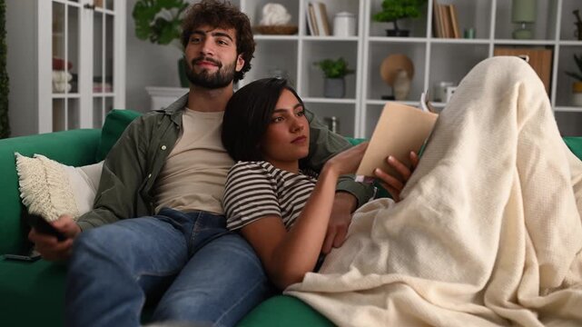 Young couple relaxing on the couch with a book and watching television in their living room