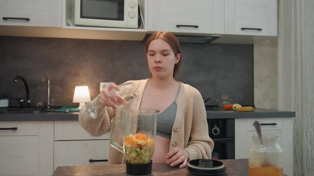 Pregnant woman in kitchen pouring water over peeled mandarins and sliced kiwi, wearing cozy cardigan and bralette, calm morning, countertop blender, preparing gentle prenatal smoothie, closeup hands,