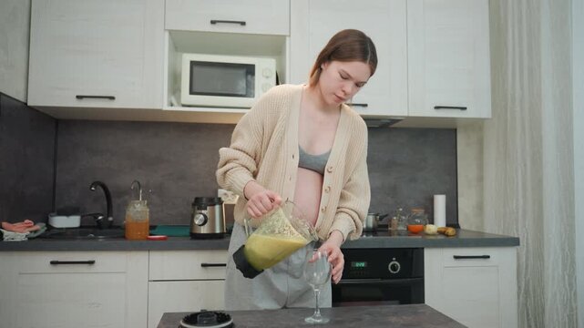 Pregnant woman pouring green smoothie into glass at counter, steady jug grip and careful pour to avoid spill, balancing glass while smiling, selfcare ritual and nourishing moment, warm cozy mood.