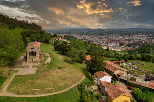 Aerial view of Santa Mar&iacute;a del Naranco overlooking Oviedo, with historic pre-Romanesque architecture, green hills and dramatic sky at sunset, Asturias, Spain.