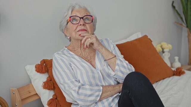 Elderly woman on bed wearing glasses and striped blouse with hand on chin and legs crossed inside a building; reflection.