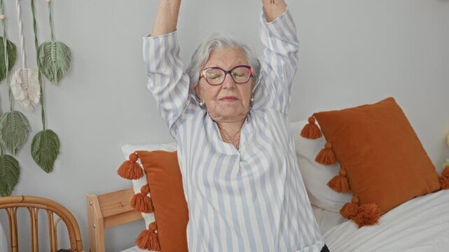 Woman stretching arms behind head on bed with orange tassel pillows and striped blouse in building, wearing glasses and eyes closed; serenity rest.