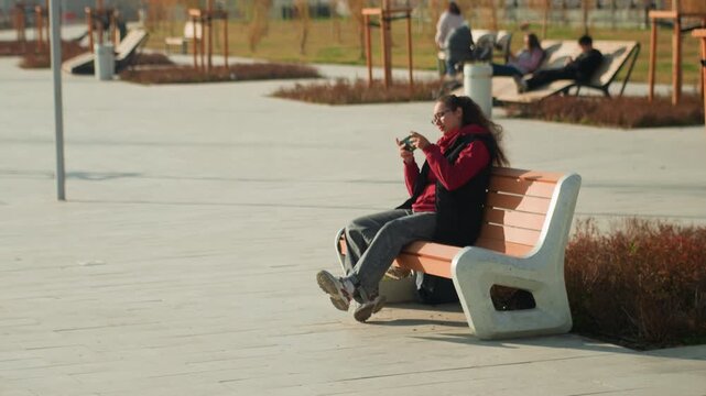 Black woman sitting on bench distressed red jacket urban park slump head in hands phone tucked away autumn chill concrete plaza passersby distant buildings muted light introspective mood anxiety