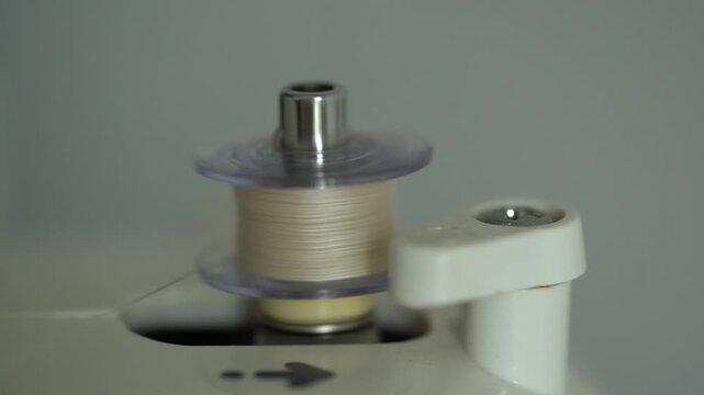 Close-up of a sewing machine bobbin almost full of beige thread continuing to wind on the bobbin winder mechanism.