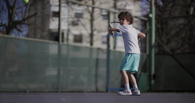 A young boy in a white shirt and blue shorts plays tennis on an outdoor court, showcasing a forehand stroke with determination.