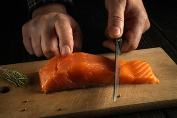 Hands hold scissors as they cut a salmon fillet on a wooden board. The setting is a kitchen with dim lighting. Fresh herbs lie nearby on the board © Віталій Б.