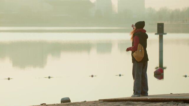 Lakeside lone person watching water, commuter pauses on foggy morning by calm urban lake, wearing red scarf and backpack, soft reflections on still surface, muted skyline in background, contemplative