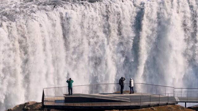 Passionate explorers dressed brightly photograph stunning waterfall Dettifoss shrouded in fog and rugged terrain