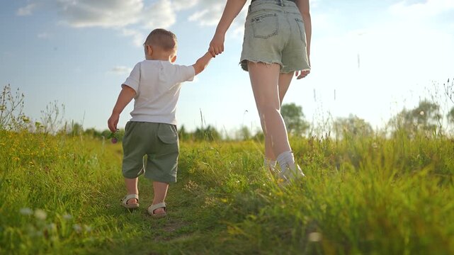 Mother and child walk holding hands on meadow path during summer. Woman walks with boy outdoors in field. Child holds mother hand while walking. Parent and son enjoy meadow time together.