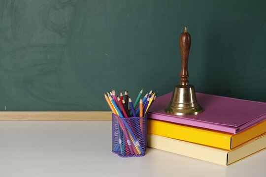 School golden bell, folders and pencils in holder on white table near green chalkboard. Space for text