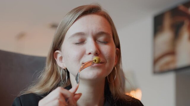 Young white woman tasting olive smiling with closed eyes holding fork cozy living room warm lamp light food blogger vibe playful sampling subtle delight slow savoring relatable lifestyle moment