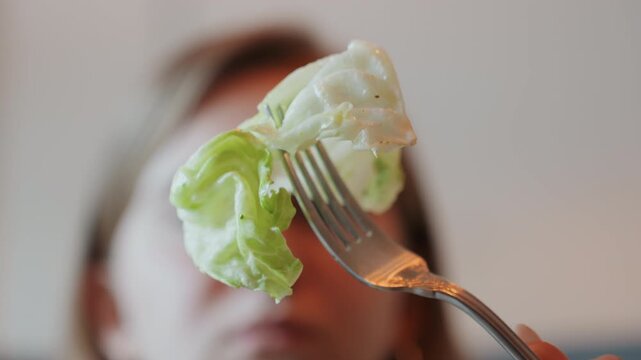 Blurry background woman prepares to bite lettuce. Closeup shot of woman holding lettuce with hesitant expression. Indoor scene capturing woman with blurred face holding lettuce on fork