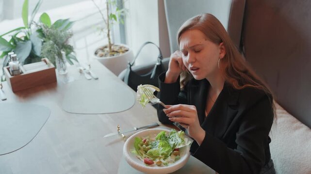 Woman in blazer waiting at lunch, urban cafe scene showing distracted professional pausing between bites, handbag at seat and bowl of greens on table, soft window light, weekday work pause with subtle