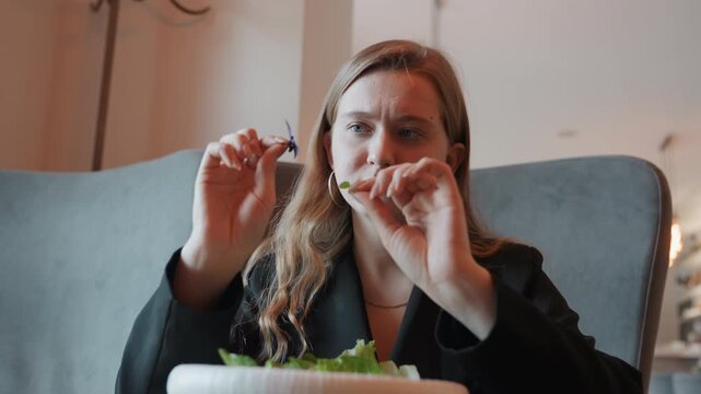 White woman examining salad leaf closely, hands and fork in frame, focused expression, blazer and hoop earrings, indoor cafe interior, picky evaluation of texture and freshness, mindful eating scene