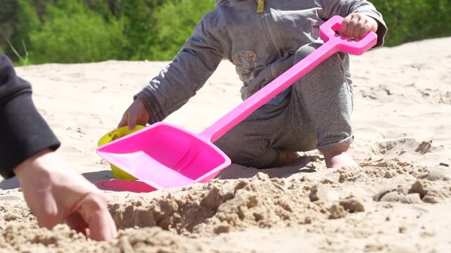Toddler playing with big pink shovel in sandbox next to adult hand shaping sand mold representing family bonding and early childhood sensory development concept