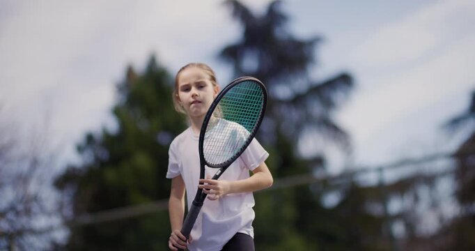 A focused young girl in a white t-shirt plays tennis, swinging her racket at a ball on a sunny outdoor court.