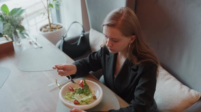 White woman testing forkful at cafe, dressed in tailored blazer, lifting forkful of greens, examining texture and dressing, focused expression, tasting salad, deciding next bite, cutlery and plate
