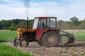 Obraz premium Old yellow tractor plowing agricultural field raising dust during spring seasonal farming work with green forest and blue sky in the background countryside