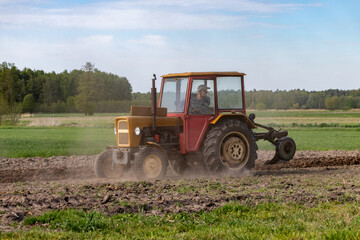 Fototapeta premium Old yellow tractor plowing agricultural field raising dust during spring seasonal farming work with green forest and blue sky in the background countryside