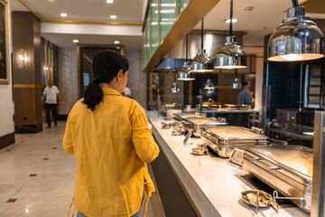 Fototapeta premium A woman with long black hair wearing a yellow jacket stands at a buffet in a hotel. The setting is modern with various food options displayed.