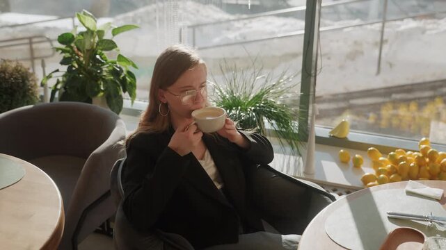 White woman gazing toward sunny window, lemons and plants on tabletop, open planner and pen beside cup, reflective smile in casual blazer and glasses, cozy interior with winter street view, leisurely