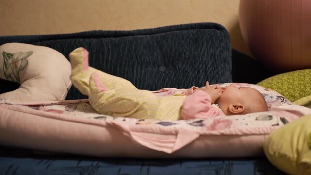 A joyful little baby, puts his hands and fingers in his mouth, tastes, plays alone with himself, has fun, examines the room. A child in a children's overalls, on a bed in a cocoon. Side view