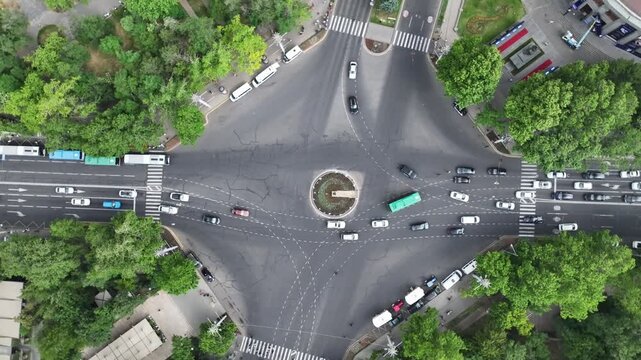 An aerial top down view of a busy city intersection with cars moving around a central roundabout surrounded by lush green trees on a bright sunny summer day.

