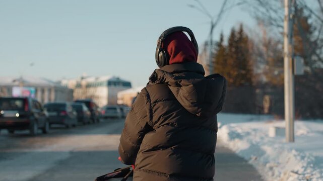Young woman walking on snowy street, adjusting headphones, puffy black jacket and maroon beanie, parked cars along sunlit suburban road, quiet morning breath visible, slow introspective pace
