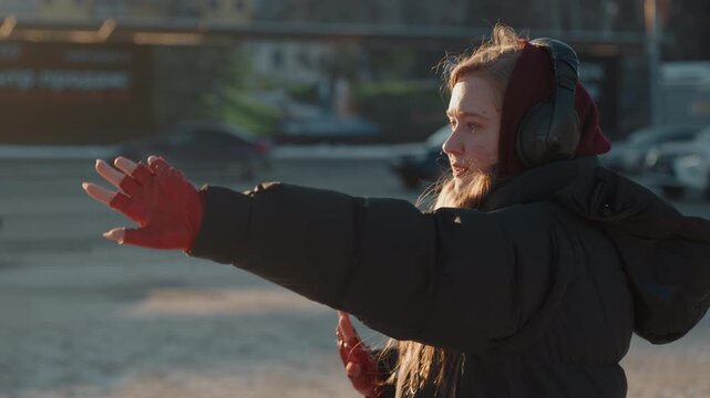 Frozen city square young woman dancing in red gloves and earmuffs, expressive hand gestures toward sunset, winter coat and steaming breath, cars and plaza background, contemplative street performer