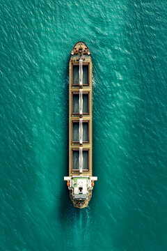 Top down aerial view of a large industrial bulk carrier or cargo ship sailing through vibrant turquoise ocean water, transporting goods and international freight