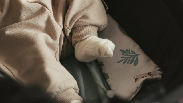 Static close up shot of a baby wearing a beige romper and white socks moving their legs while lying in a stroller with a patterned pillow.