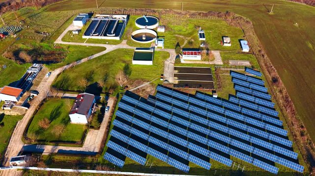 Aerial view of industrial water treatment plant and solar panels. Drone shot of water purification utility infrastructure with green fields and photovoltaic solar array panel rows.