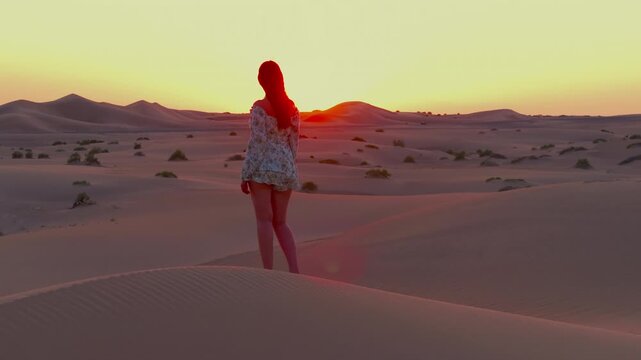A young women with long hair wearing a summer dress and walking barefoot across vast sand dunes